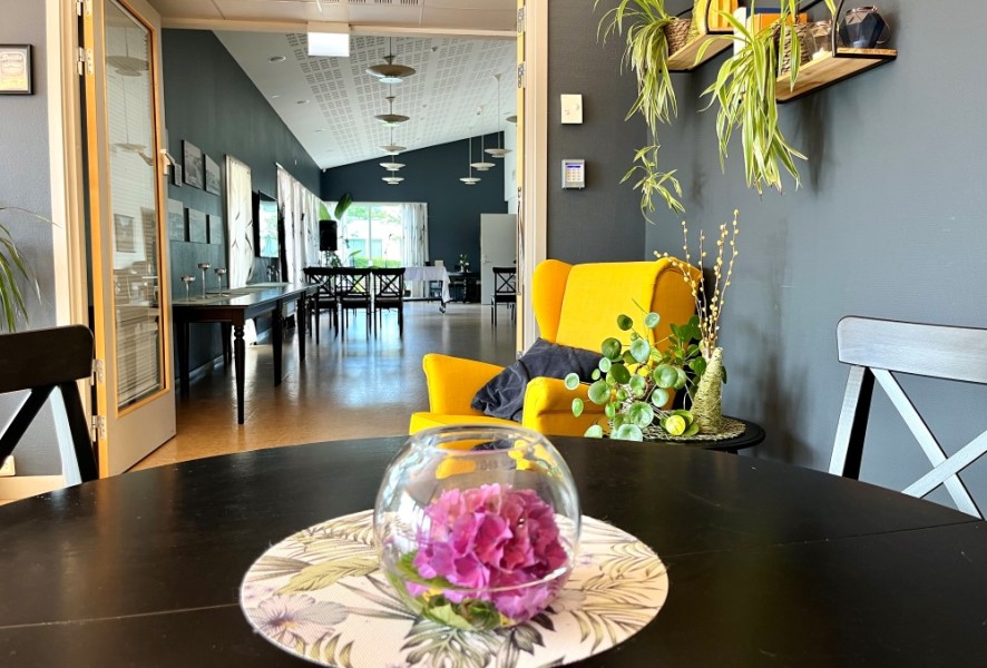 Bildet viser deler av et sort bord i kafeen med en blomst i en vase. I bakgrunnen ser vi en gul lenestol og rommet bak, som er aktivitetsrommet. 