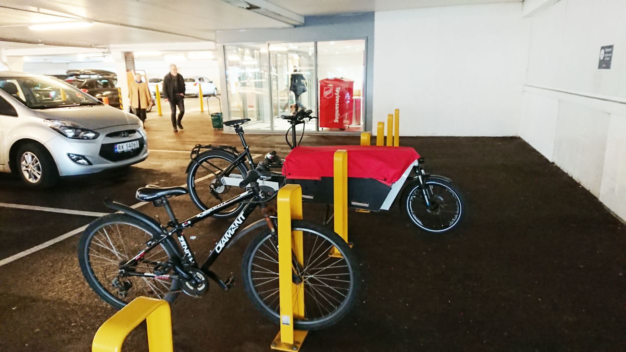 En lastesykkel og en vanlig sykkel står parkert i stativer i et parkeringshus til et kjøpesenter i forbindelse med bilparkeringen.
