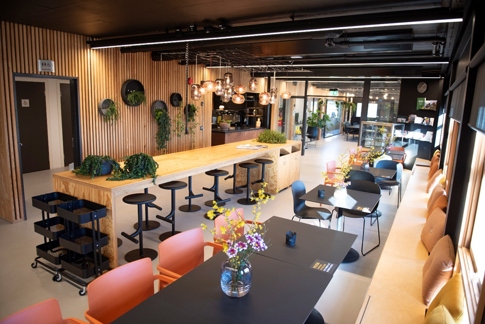 Oversiktsbilde fra kafeområdet. Et høyt langbord med ca 10 barstoler, sitteplasser i benk langs vindu, grupper med bord og stoler.