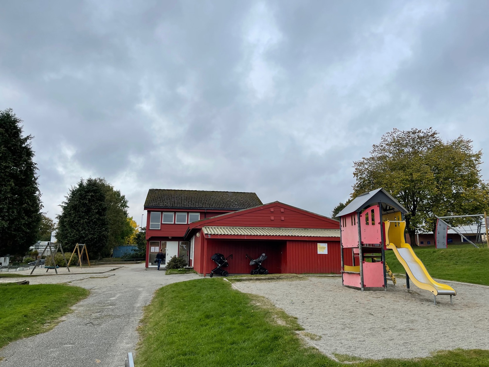 Overblikk over veden åpen barnehage. Rød barnehage med plen rundt. Noe grus og asfalt i nærheten. Foran barnehagen står et lekestativ.