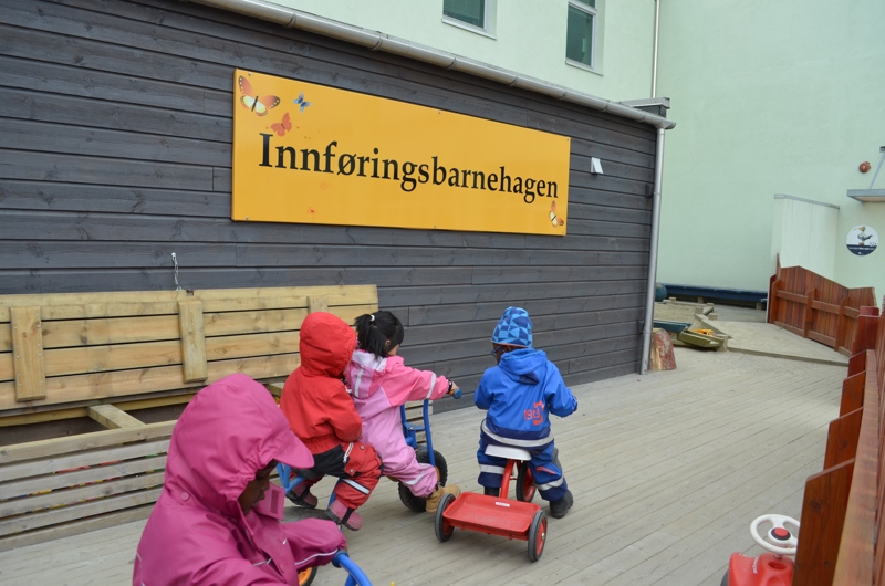 Utendørs bilde av fire barn som leker på små sykler og et trehjulsykkel på en treplatting foran et brunt bygg. På veggen henger et stort gult skilt med teksten "Innføringsbarnehagen".