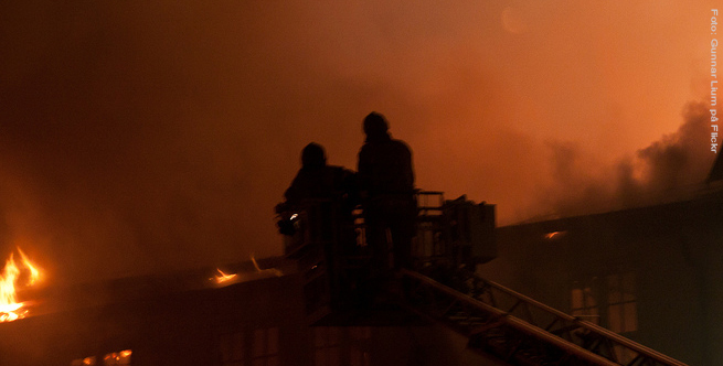 Brannmenn i korga til en brannbil, midt i røyken over et hus som brenner i mørket