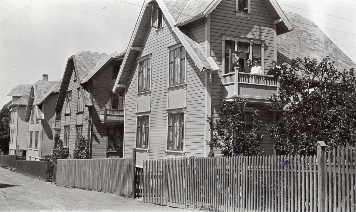 Svart-hvitt-bilde som viser fire hus i gamle dager, med tidsriktige vinduer. Fire personer står på balkongen på det nærmeste huset.