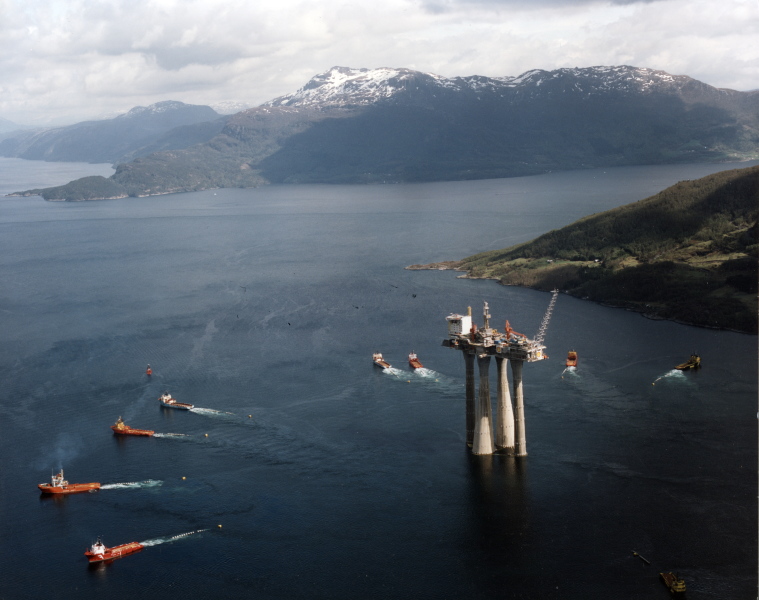 En enorm plattform slepes av åtte båter, gjennom norsk fjordlandskap.