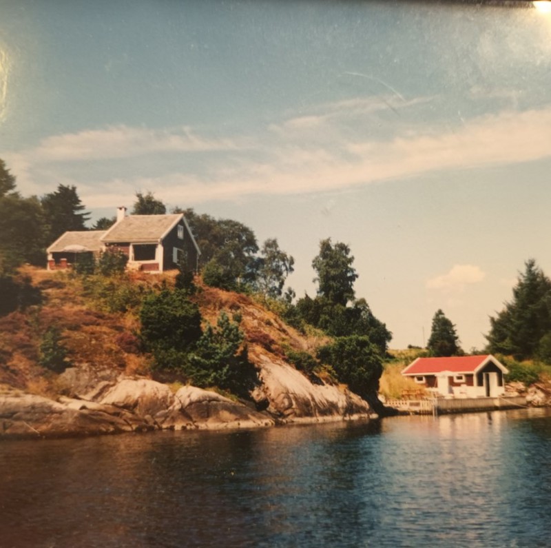 En liten, etterkrigsmodernistisk hytte av to saltaksvolum oppå et bratt svaberg, med naust og brygge nedenfor.