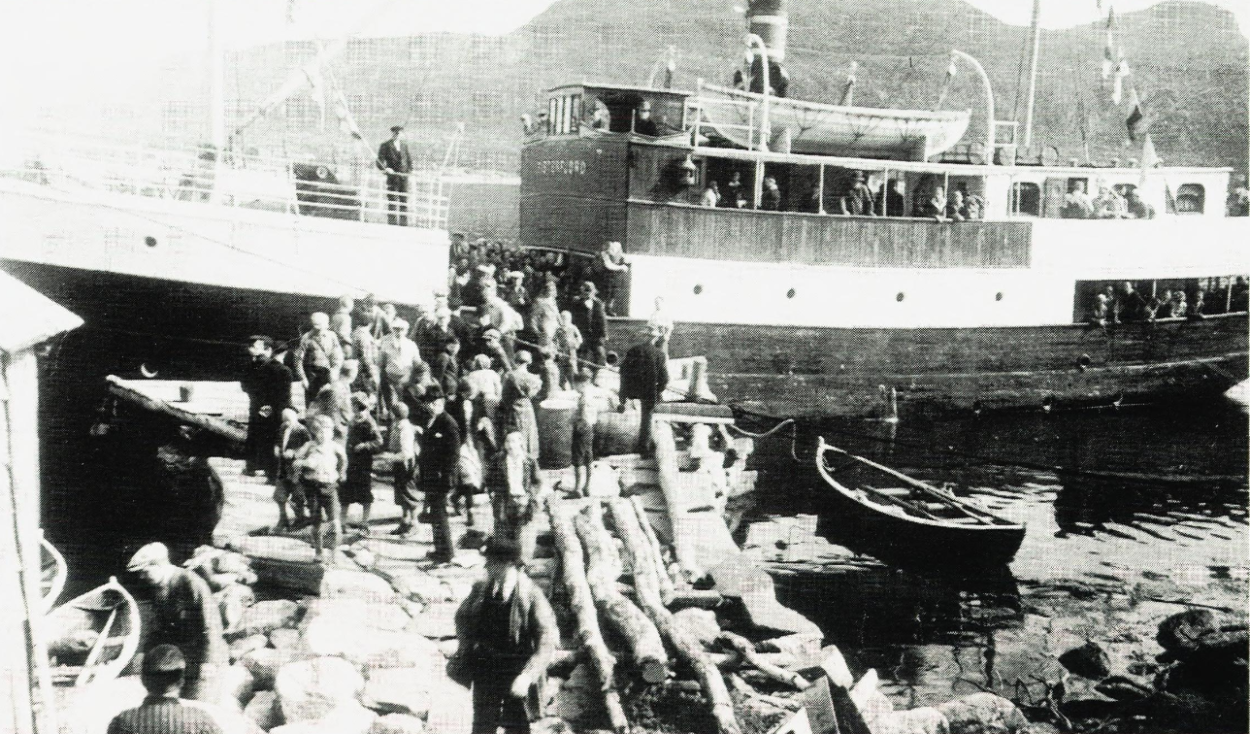 Svart-hvittbilde av dampskip ved liten kai eller brygge. Mange folk på brygga, og en færing i sjøen innenfor dampskipet.