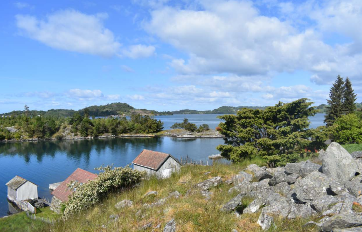 Gamle naust ved en rolig, blå fjord, med flere øyer og holmer. Nærmest, i bakken over naustene, ligger en steinrøys.