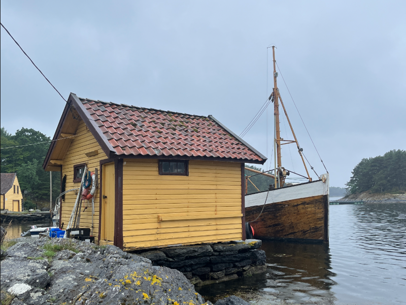Liten, gul sjøbod og tradisjonell fiskeskøyte ved kaien en regnværsdag om sommeren.