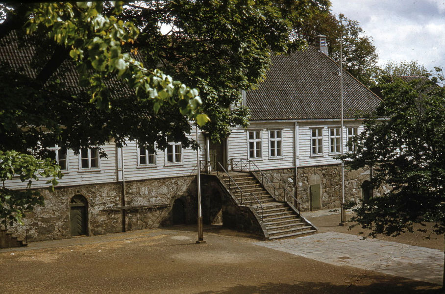 Hovedbygningen på Kongsgård skole, sett delvis gjennom løvverket til trær. Skolegården er tom, ingen flagg eller vimpler på de to flaggstengene på hver side av steintrappa.