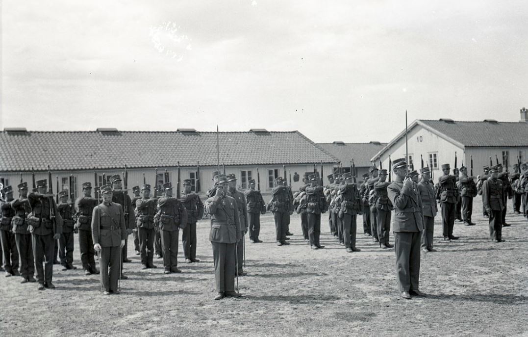 Et svart-hvittbilde av soldater oppstilt i Madlaleiren. Brakker bak soldatene.