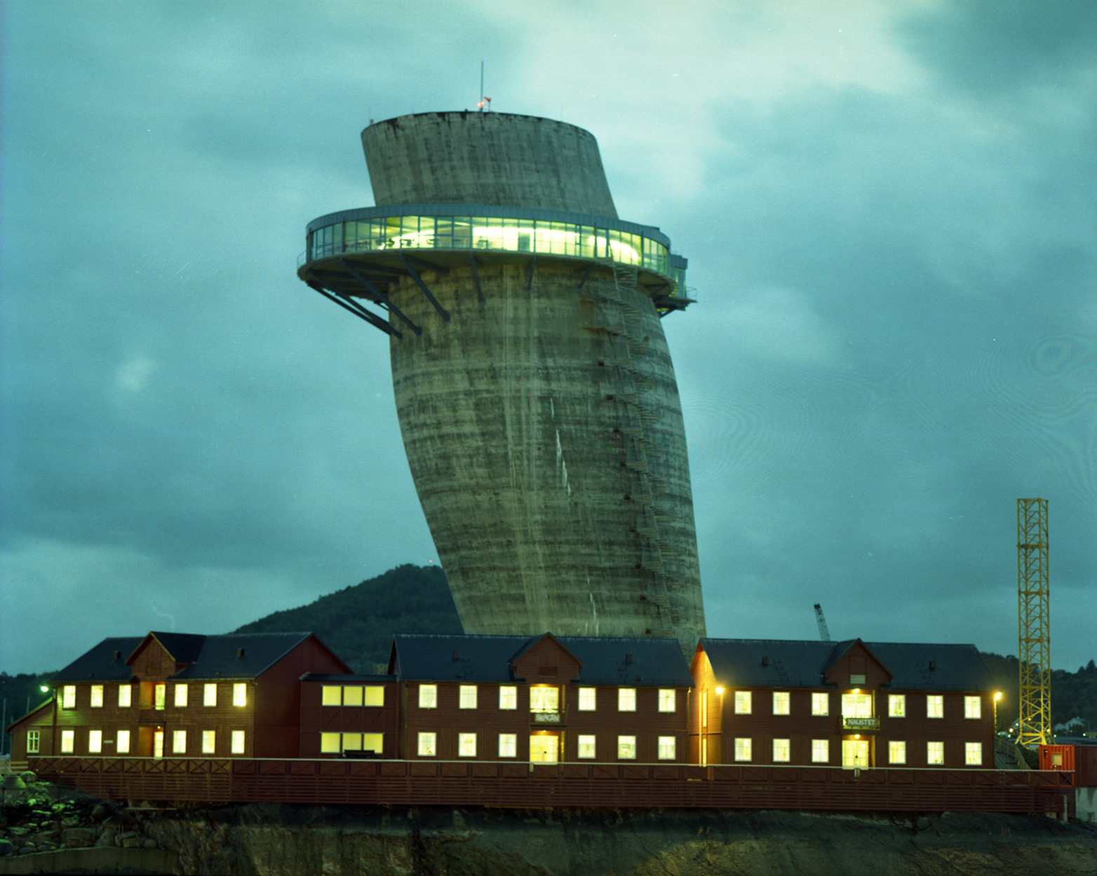 Skråtårnet i Jåttåvågen fotografert i skumringslys, med Jåttånuten i bakgrunnen og saltaksbygninger av sjøhustype i forkant. Det er lys i alle vinduer, også i den utkragede etasjen høyt oppe i Skråtårnet.