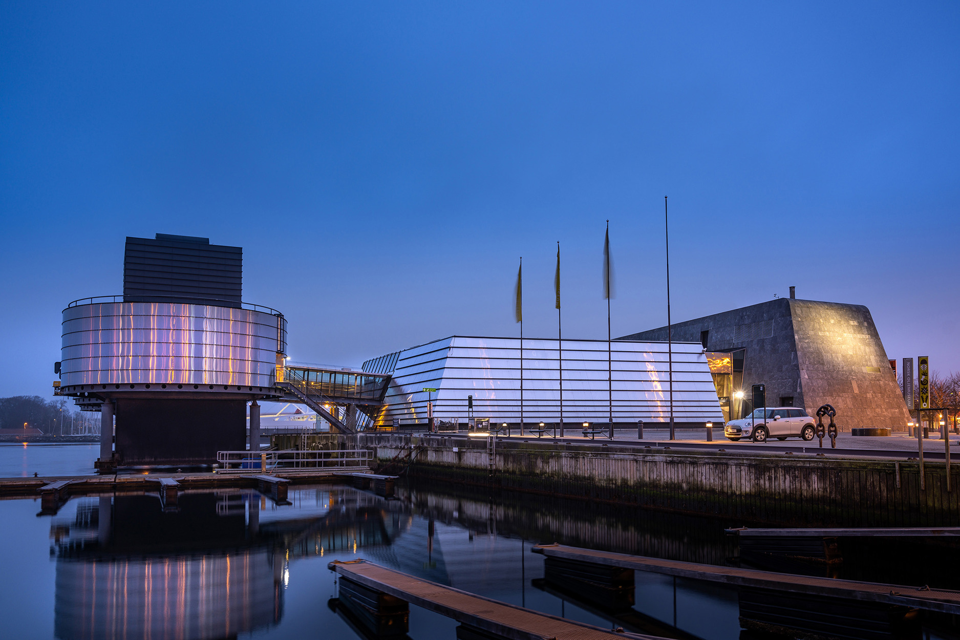 Norsk Oljemuseum sett fra Blå Promenade ved Børevika, fotografert i skumringslys en kveld med klarvær. Fjorden ligger blikkstille, og speiler oljemuseet, som igjen speiler byens lys i den nærmeste sylinderplattformen som står i sjøen.