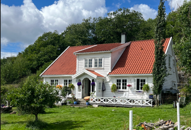 Hvitt våningshus med smårutete vinduer i grønne omgivelser.