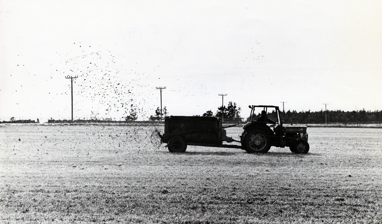 Et flatt jorde med en traktor med gjødselhenger. Både maskinene og gjødselen er i tydelig silhuett mot den lyse himmelen og marka.