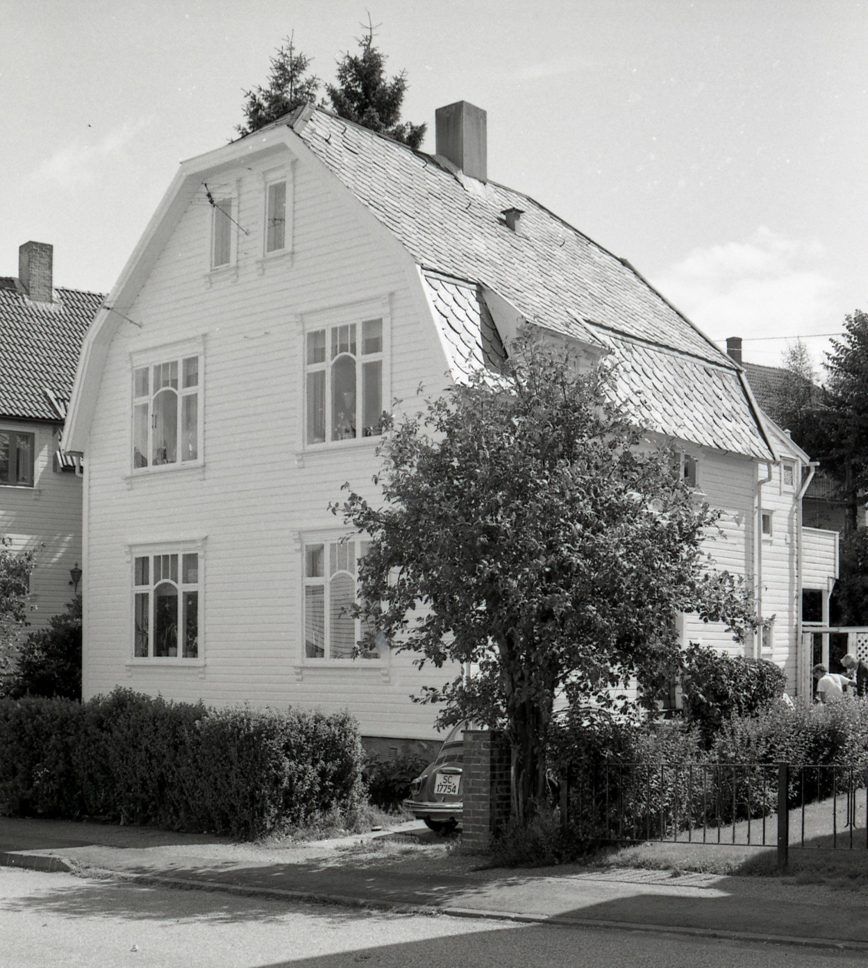 Hvitmalt tomannsbolig med hekk mot fortauet, og en VW boble parkert i innkjørselen.