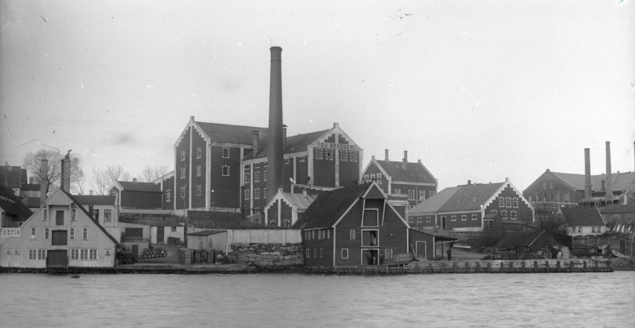 Svart-hvittbilde av Tou bryggeri sett fra sjøen. Andre bygninger i bildet er sjøhus og fabrikker med høye piper, og noen små bolighus.