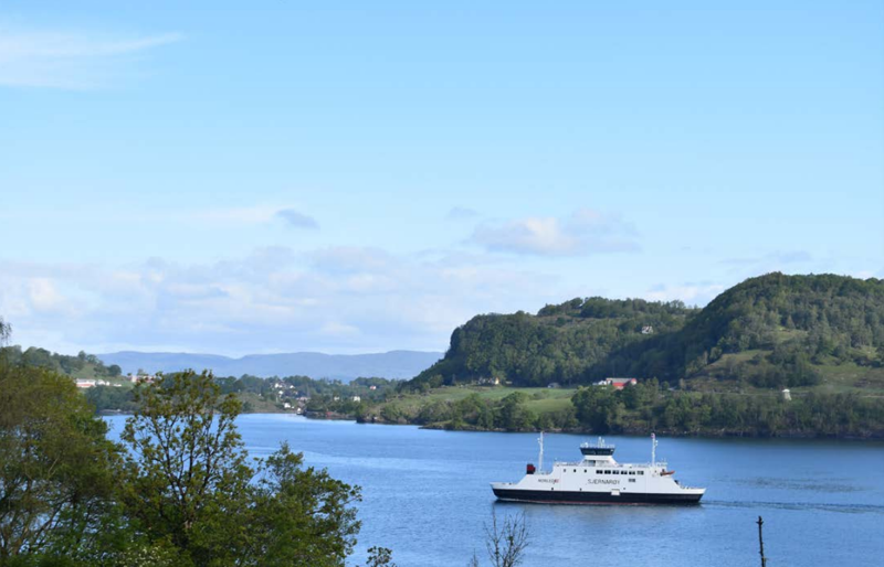 Ferga Sjernarøy på ein blå fjord, under blå himmel, med grøne, knausete øyer og holmar, med fjell lengst bak.