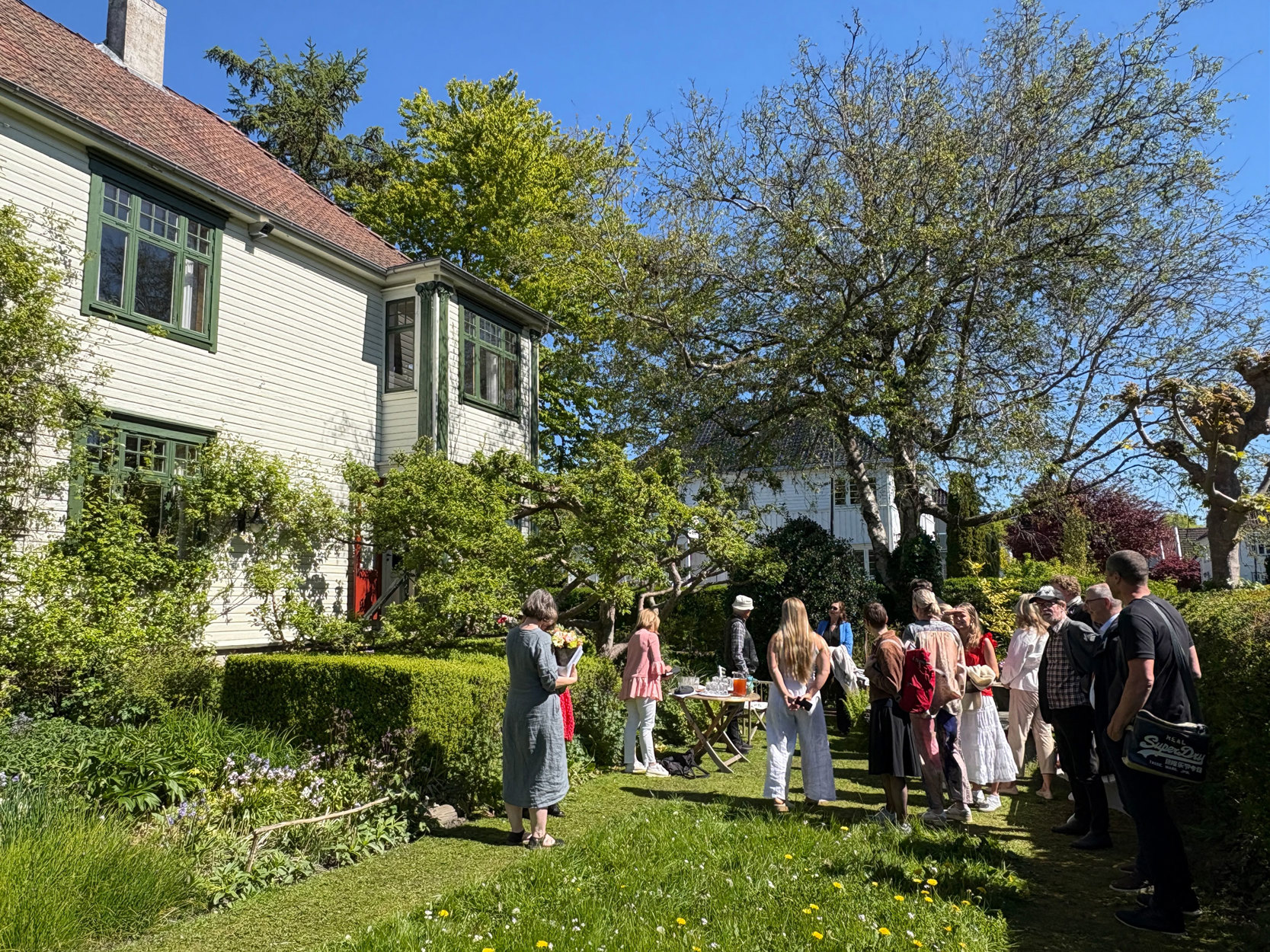 Masse folk i hagen foran huset. En del av plenen er uklippet blomstereng.