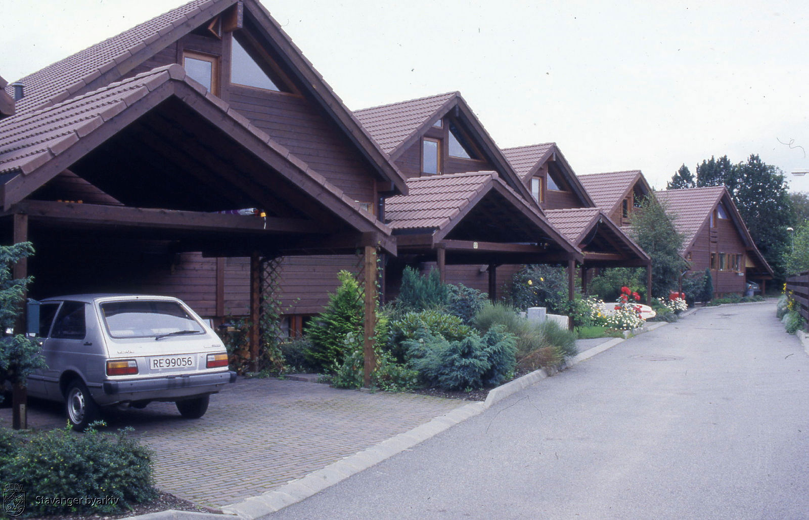 Hus på rekke og rad, med hver sin carport foran huset og mye vegetasjon langs gaten. Eldre bil står i en av carportene.
