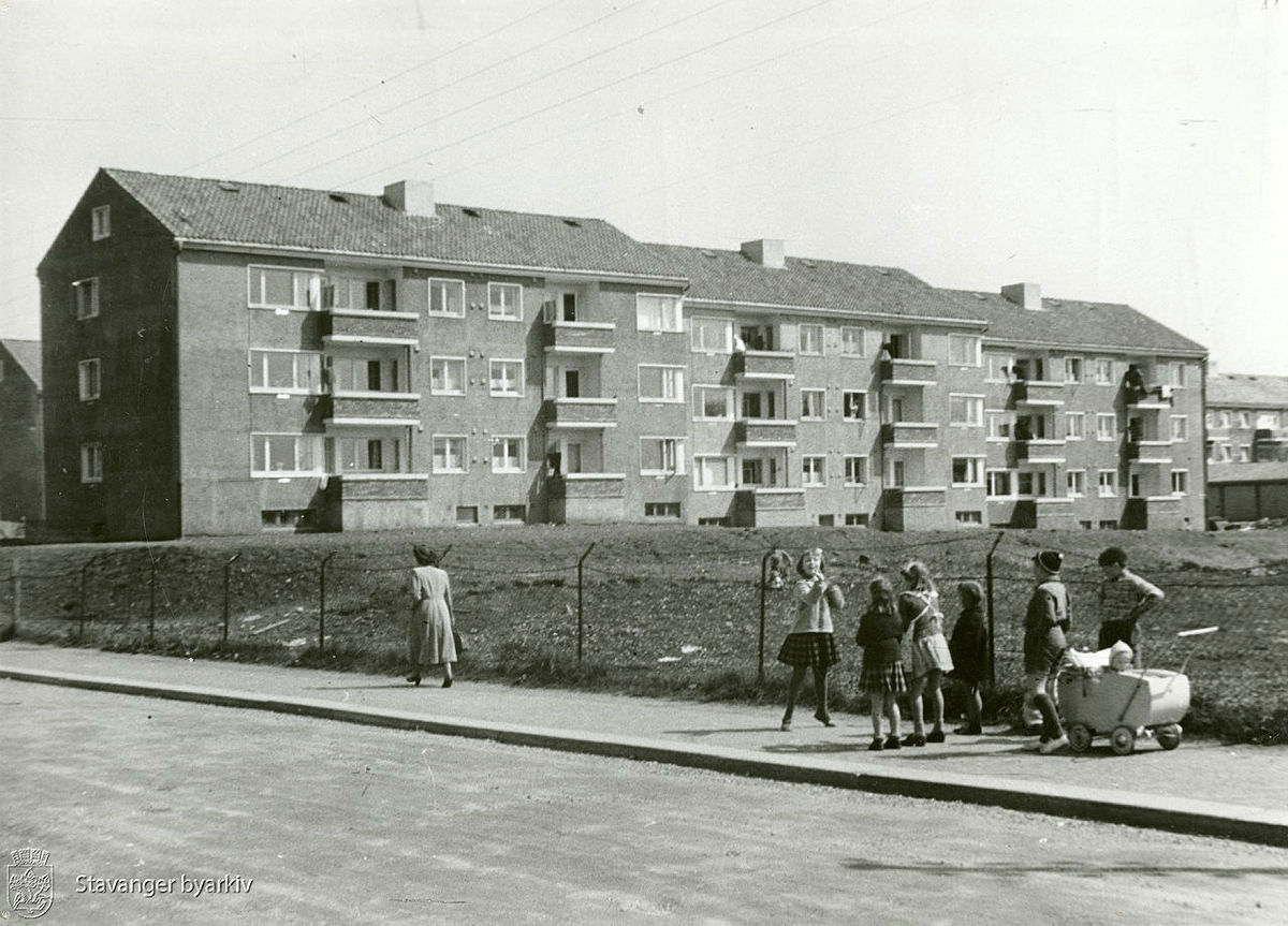 Svart/hvitt-bilde av barn som leker med bl.a. dukkevogn i forgrunnen. Boligblokker i bakgrunnen.
