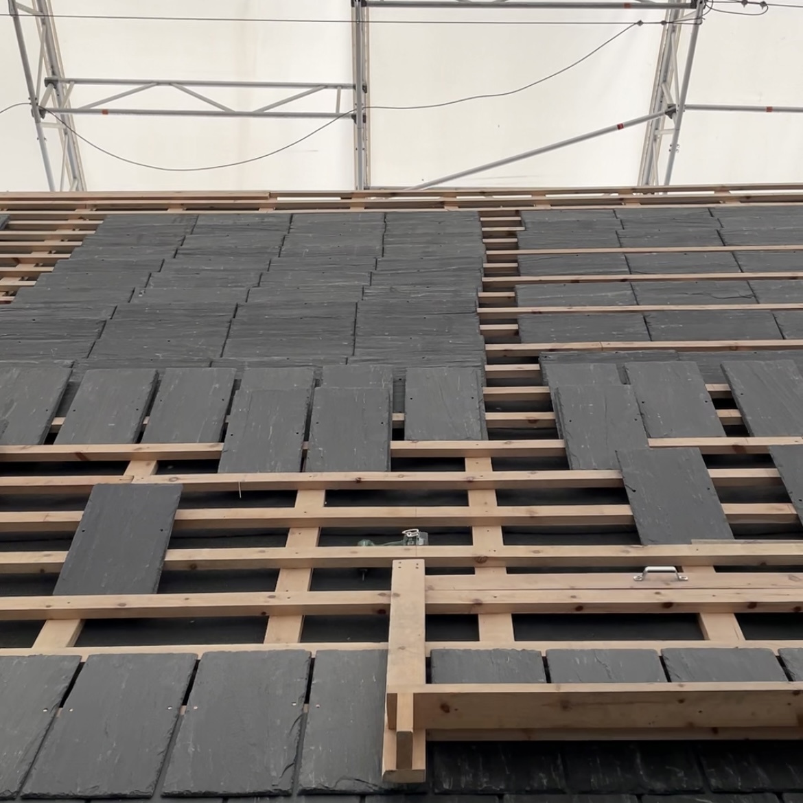 Nærbilde av arbeid med skifterstein på domkirketaket. Firkanta skifterplater der delvis lagt, delvis klare for legging, på nye lekter. Over taket skimtes den hvite presenningen som dekker hele kirken.