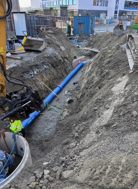 Nye rør og ledninger og en kum ligger i en lang grøft i ei gate. Anleggsmaskiner ved grøfta.