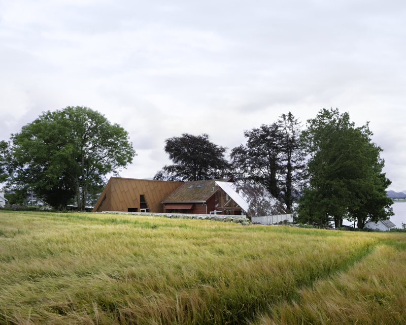 Et tun med tre bygg, med en kornåker i forkant. bygget til venstre har fasade av corténplater, huset i midten er et eldre hus, mens bygningen til høyre er en pergola av glass. 