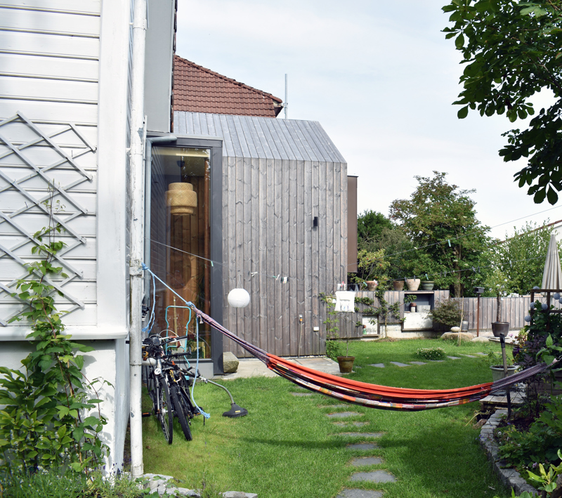 Et uterom ved en eldre bolig som har fått et moderne tilbygg. Vi ser en plen med hellebelagt sti, sykler lent inntli husvegg og en hengekøye midt i bildet.
