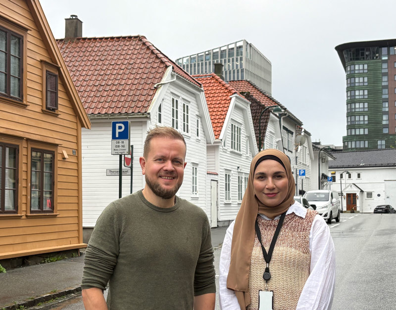 En mann og en kvinne står ute i byen og ser mot kamera. Vi ser en rekke med eldre trehus i bakgrunnen. 