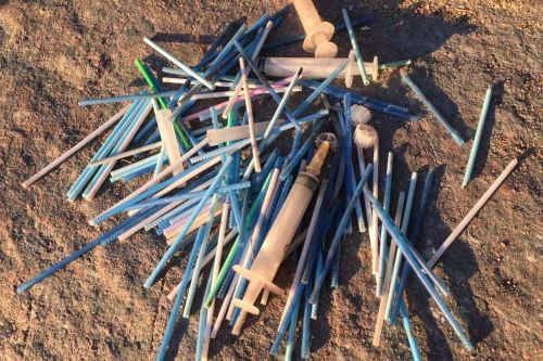 Nærbilde av mange q-tips og noe sprøyter som liggeri en haug på en sandstrand.