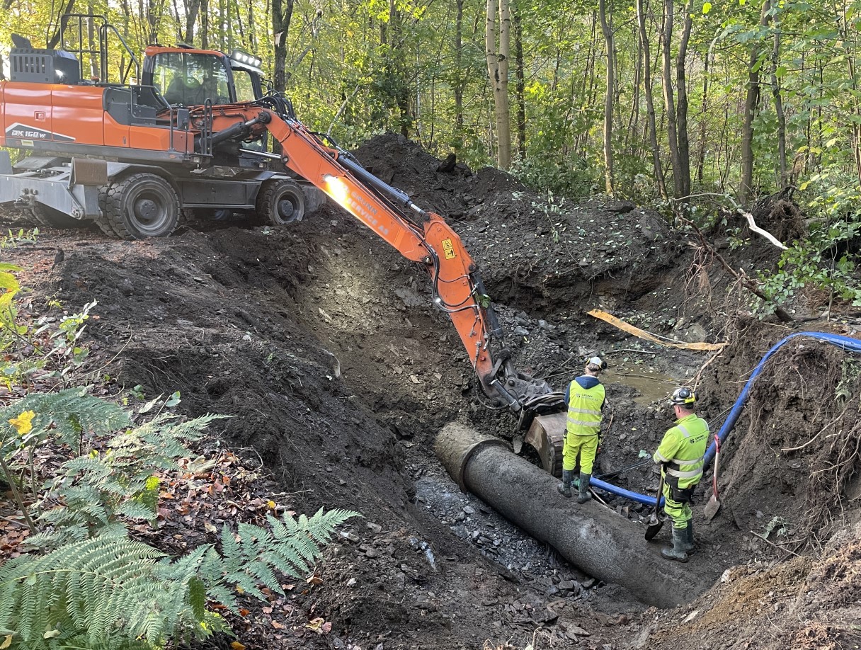Gravemaskin og arbeidere jobber med vannrør i et dypt hull i bakken.