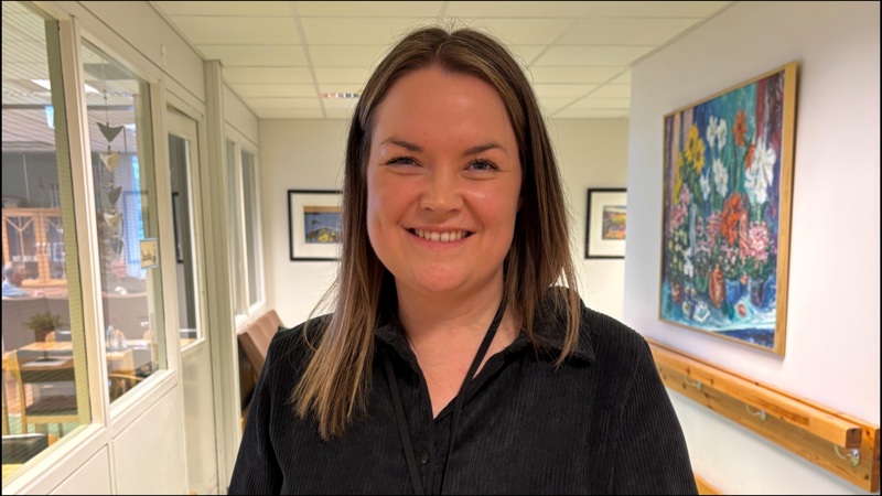 Kristine Østerli står smilende i gangen på Blidensol sykehjem. Hun har brunt halvlangt hår med midtskill og en mørk bluse.