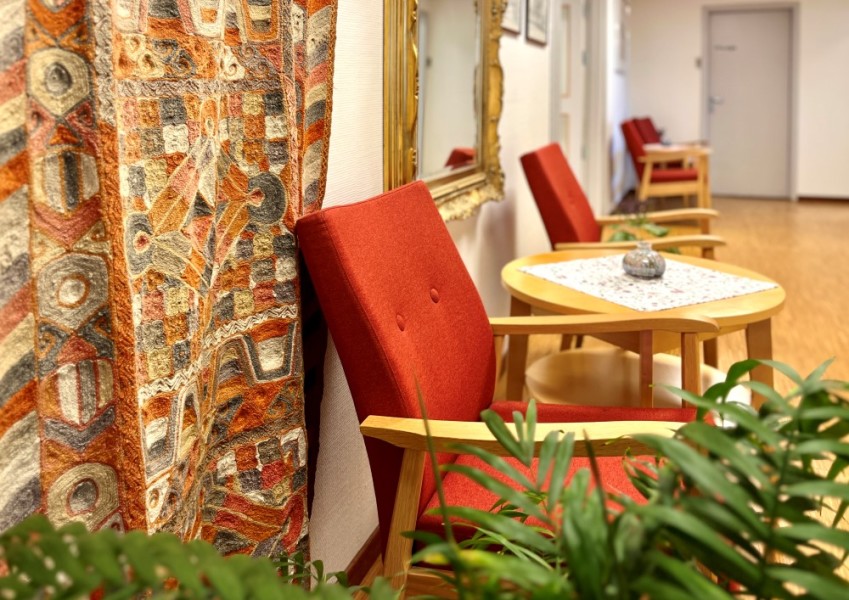 Vi ser røde stoler med bruke armlener i tre som står i gangen. Bak henger det store veggteppe i rødfarger. Det er pyntet med planter rundt.