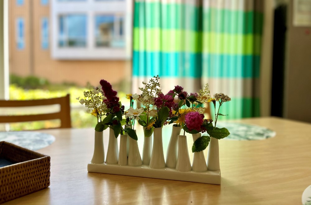 Vi ser en hvit vase med masse små sommerblomster som står på et bord.