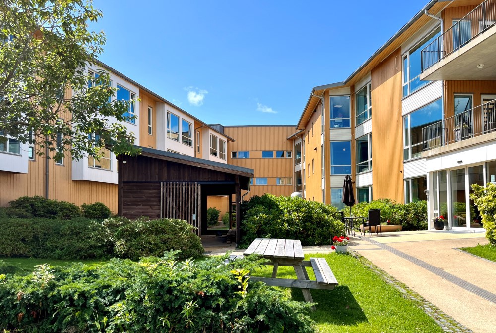 Vi ser et bildet av sykehjemmet fra utsiden, med hage og gulmalt panel.