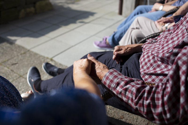 To eldre sitter på en benk og holder hverandre i hendene. Bildet er tatt skrått ovenfra, slik at ansiktene ikke synes.