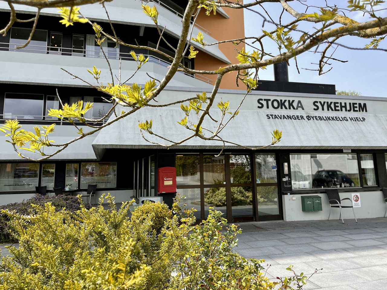 Vi ser inngangspartiet til Stokka sykehjem. Bygget er stort, grått, brunt og orange farge. Et tre med grønne vårbladet på greinene står foran inngangen.