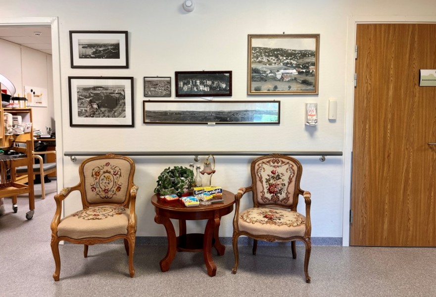Bildet viser en gang på sykehjemmet, hvor det står to gamle flotte trestoler med et lite bord mellom seg. På veggene henger en gruppe gamle sort hvitt bilder av Stavanger, og folk.
