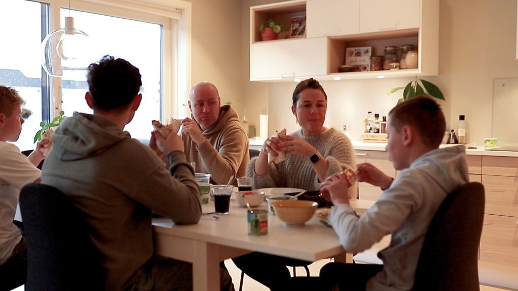 En familie med to voksne og tre barn sitter rundt et bord og spiser.