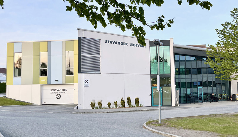 Vi ser Stavanger legevakt fra utsiden. Et hvitt betongbygg, delt inn med glassvinduer og dekorplater i gult på den ene veggen. 