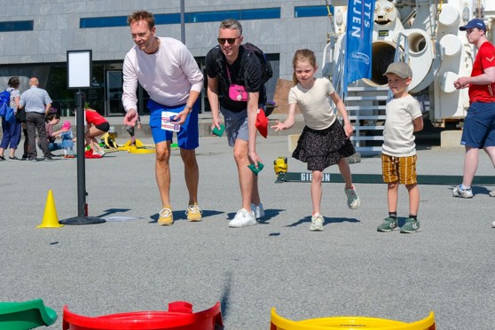 Vi ser to menn og to barn som står på en rekke. De kaster ball, og prøver å treffe et hull i en konkurranse.