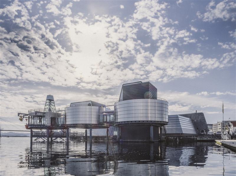 Det norske oljemuseet i Stavanger sett fra sjøsiden, med moderne, sirkulære bygninger i metall, og speilbilde i vannet under en delvis skyet himmel.