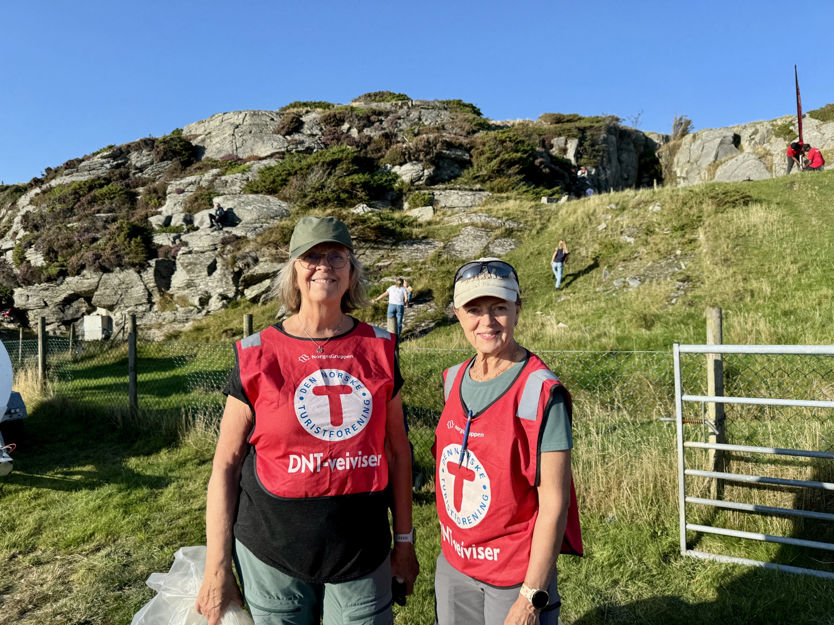 To frivillige kvinner i røde vester smiler til kamera.