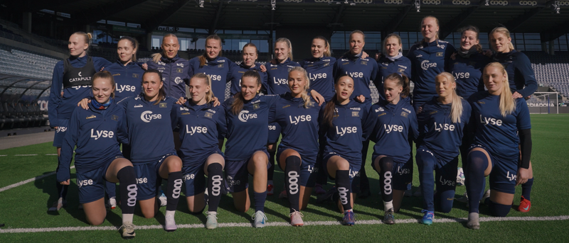 Kvinnelag ikledd vikingdrakt står på banen og synger inn i kamera. 