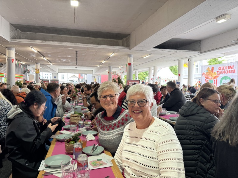 To voksne kvinner smiler til kamera. Sitter på langbord. 