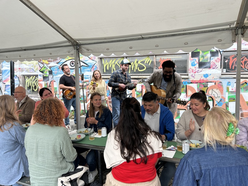 Band spiller musikk mens mennesker langs langbord spiser mat. 