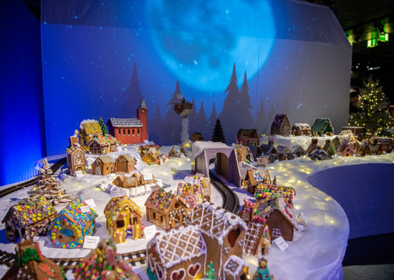 En utstilling av en pepperkakeby med mange små hus dekorert med fargerik glasur og godteri. Husene står på et hvitt underlag som imiterer snø, og et togspor slynger seg gjennom byen med en tunnel i midten. I bakgrunnen er det en stor blå projeksjon av en måne og silhuetter av grantrær, som gir en vinterlig stemning.