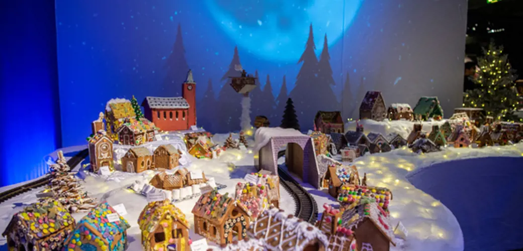 Foto: Rune Egenes/Norsk Oljemuseum En utstilling av en pepperkakeby med mange små hus dekorert med fargerik glasur og godteri. Husene står på et hvitt underlag som imiterer snø, og et togspor slynger seg gjennom byen med en tunnel i midten. I bakgrunnen er det en stor blå projeksjon av en måne og silhuetter av grantrær, som gir en vinterlig stemning.