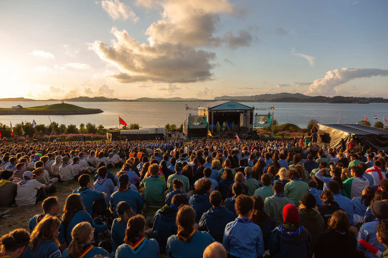 Mange tusen speidere sitter på gressplenen og lytter til en konsert. Solnedgang og sjø i bakgrunnen.