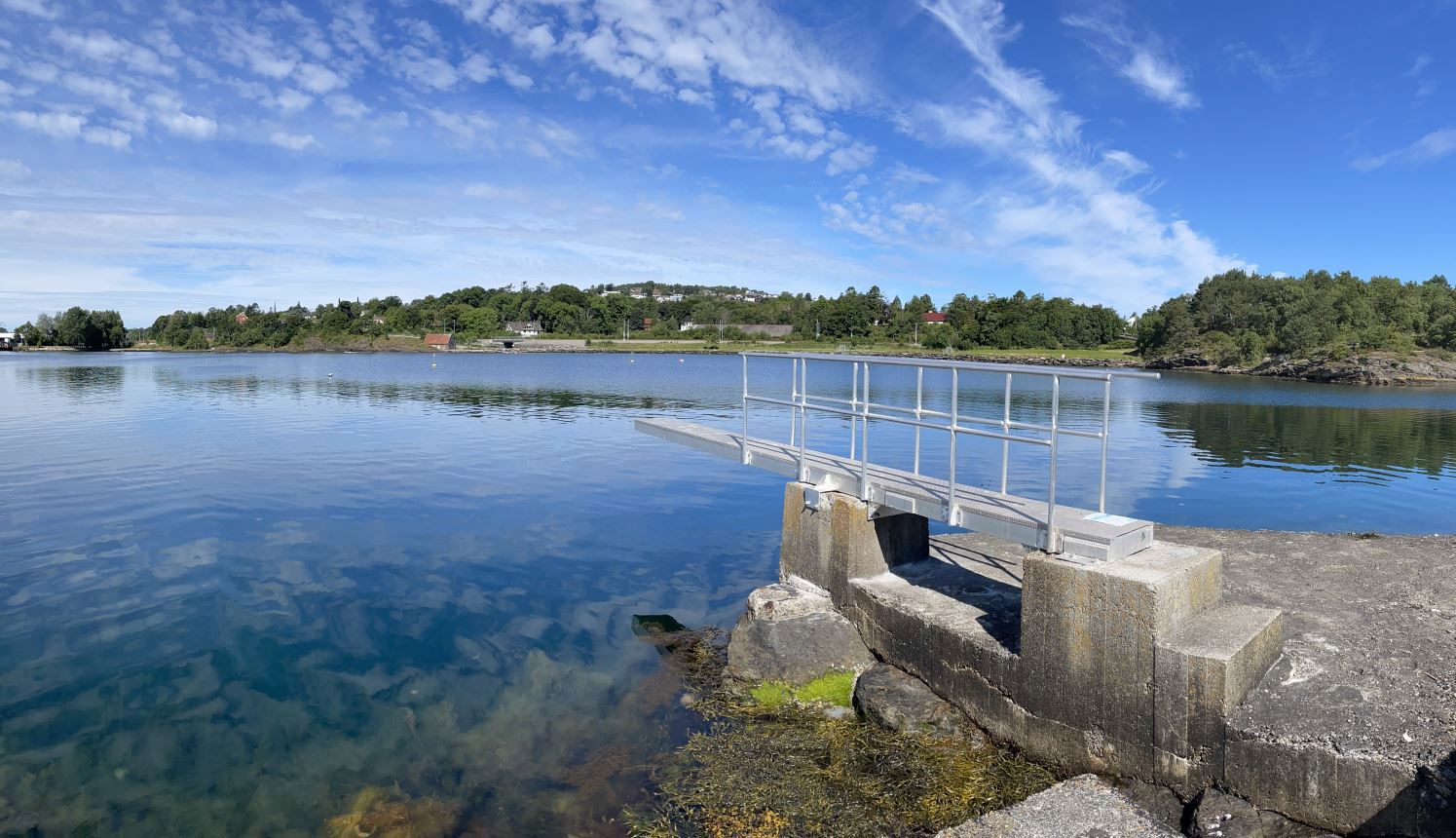 Stupebrettet er satt opp sør på Vaulen badeplass. Stupebrett i stål på betongfundament. Himmel med hvite skyer speiler seg i mørkeblå sjø.